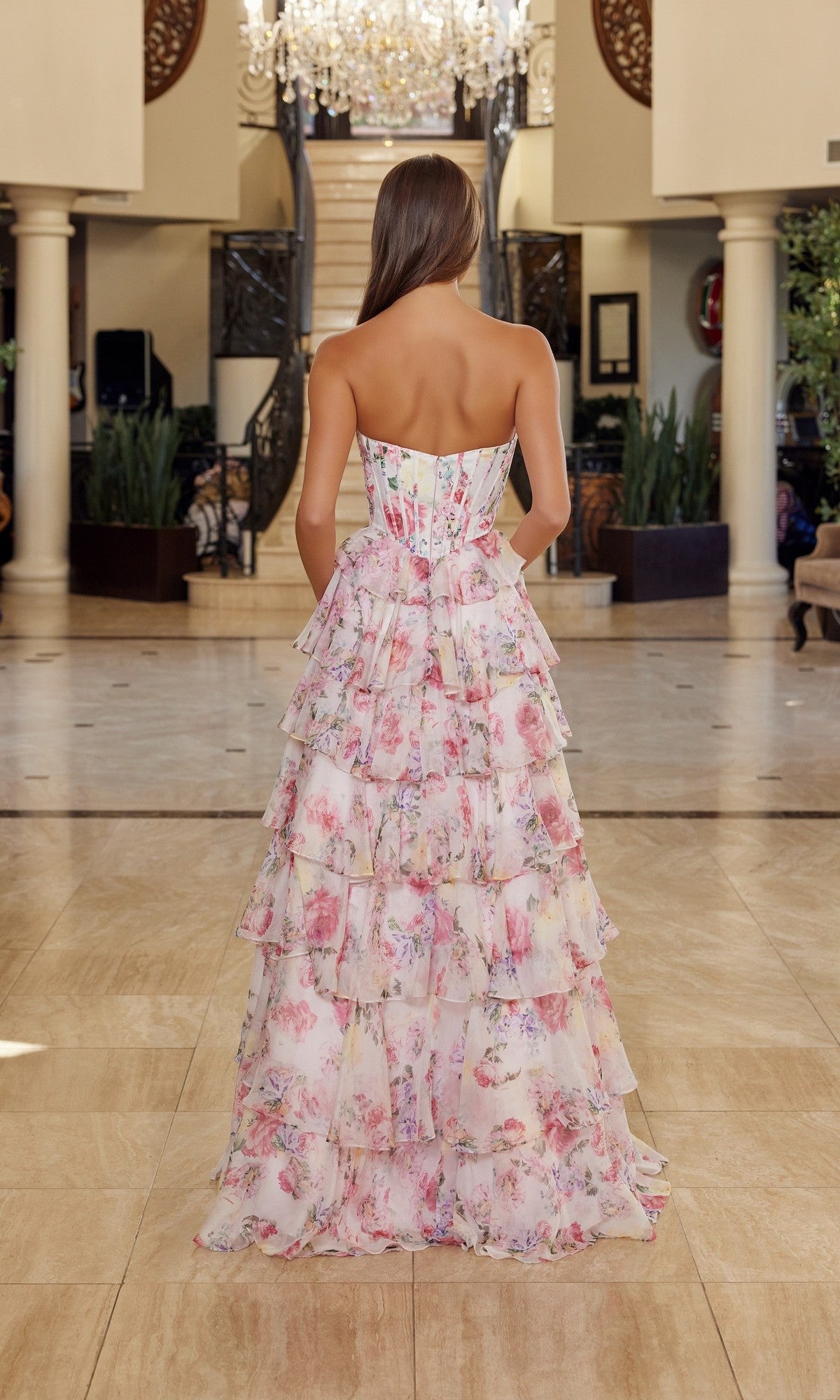 A woman stands indoors, facing away from the camera, wearing the Nox Anabel Q1779 long prom dress. The floral strapless gown features chiffon tiered ruffles, with a chandelier and grand staircase in the background.